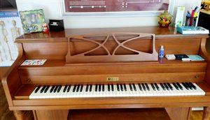 [Image of a LeSage upright piano in medium brown wood. There are ornaments from students, a sticker sheet, and flashcards on top of the piano.]